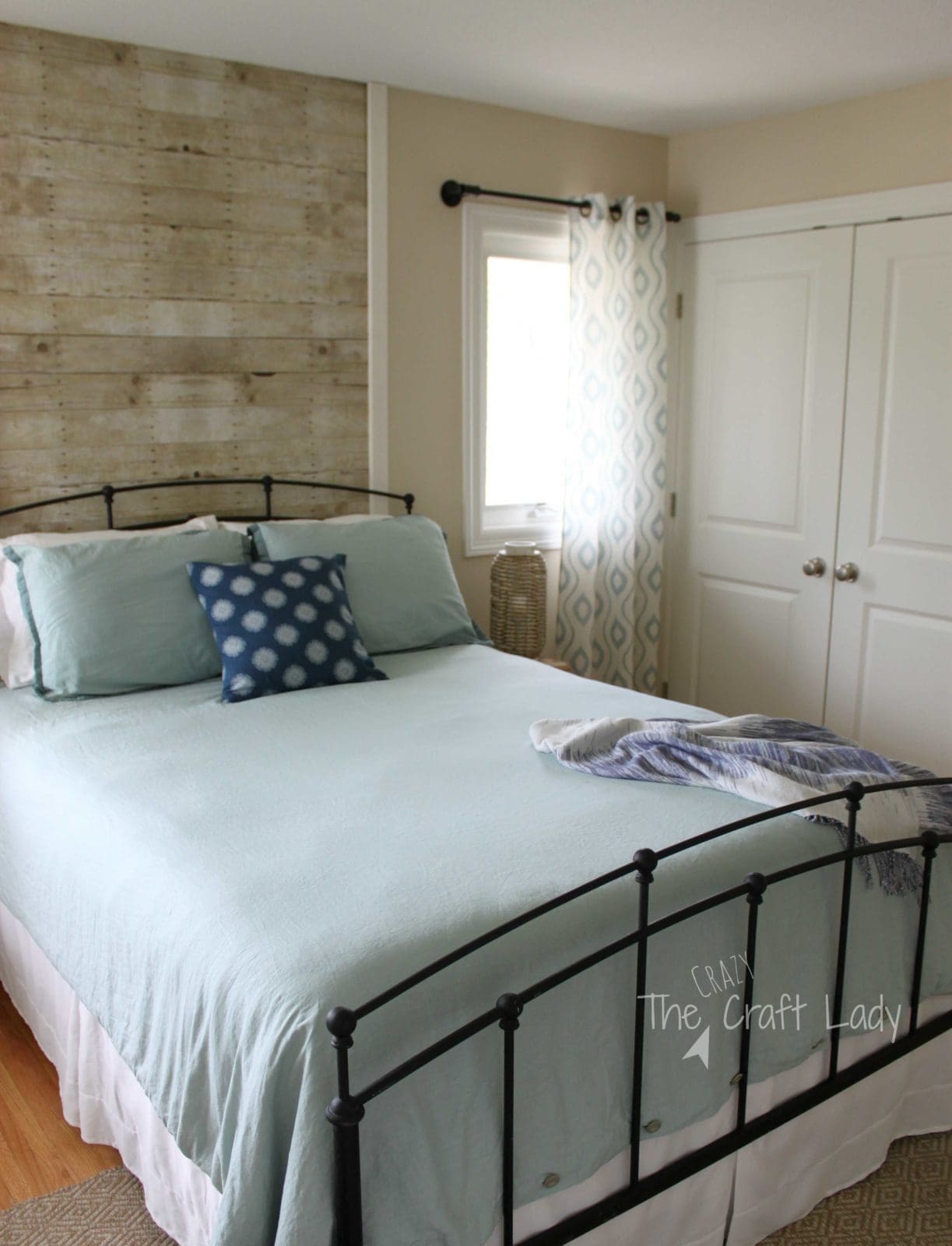 A rental-friendly plank wall in light wood behind a black frame bed with a white comforter. A window with light curtains and a double door white closet are to the right. 