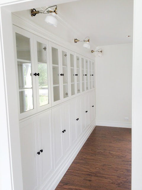 A row of white IKEA Borgsjo bookcases have glass doors on the top half and long cabinet doors on the bottom. This DIY IKEA hack provides gorgeous built-in shelves. 