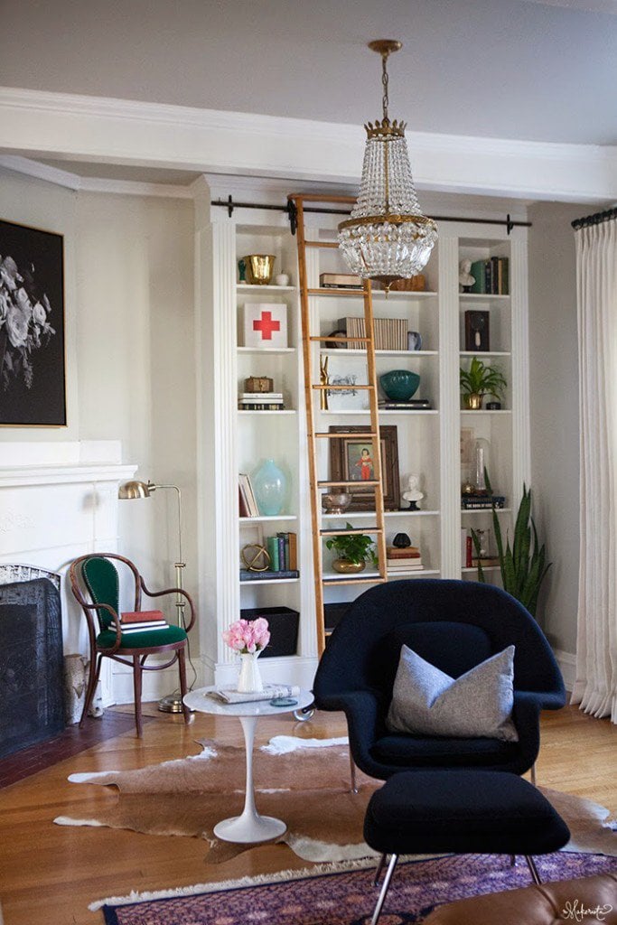 A living room with a fireplace, simple chairs, and a DIY IKEA hack made from Billy bookcases that stand floor-to-ceiling and include a ladder to reach the higher shelves. 