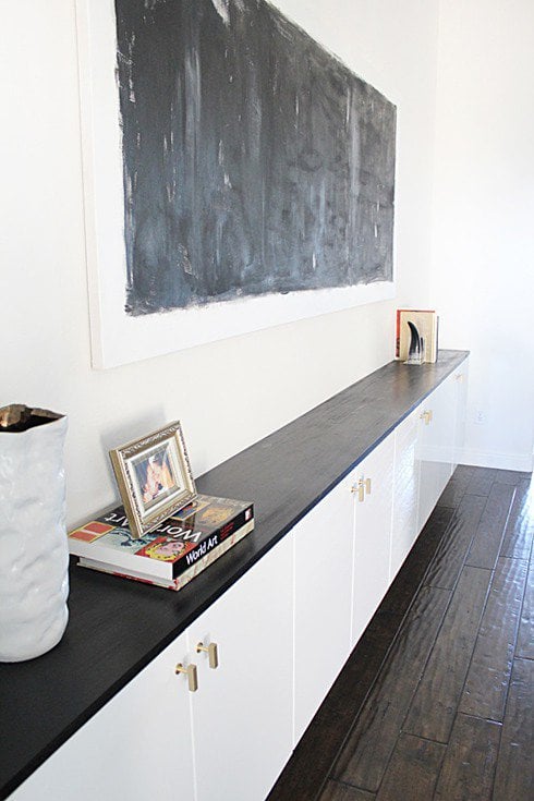 Kitchen cabinets have been altered to line a living room wall as floating cabinets. Abstract art hangs above this dark wood and white DIY IKEA hack.  