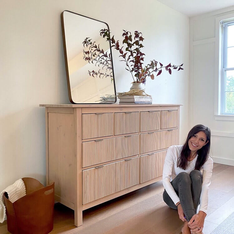 A smiling woman sits on a wooden floor next to a fluted Hemnes dresser altered using DIY IKEA hacks.