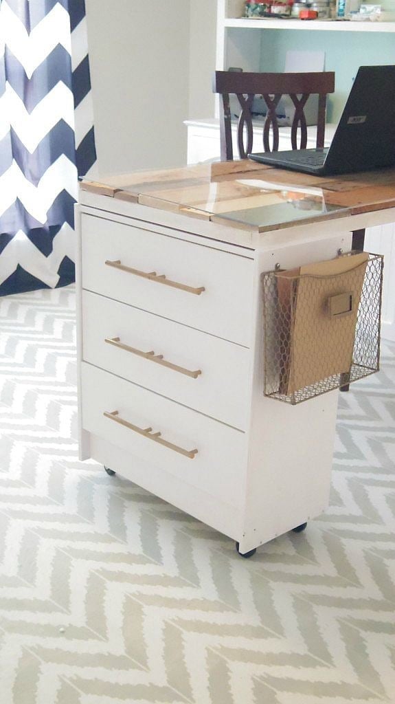 A Rast desk from IKEA has been upgraded with off-white paint, gold casters and a wood and glass desktop. Blue and white chevron curtains hang behind the desk, and white shelves line the wall. 