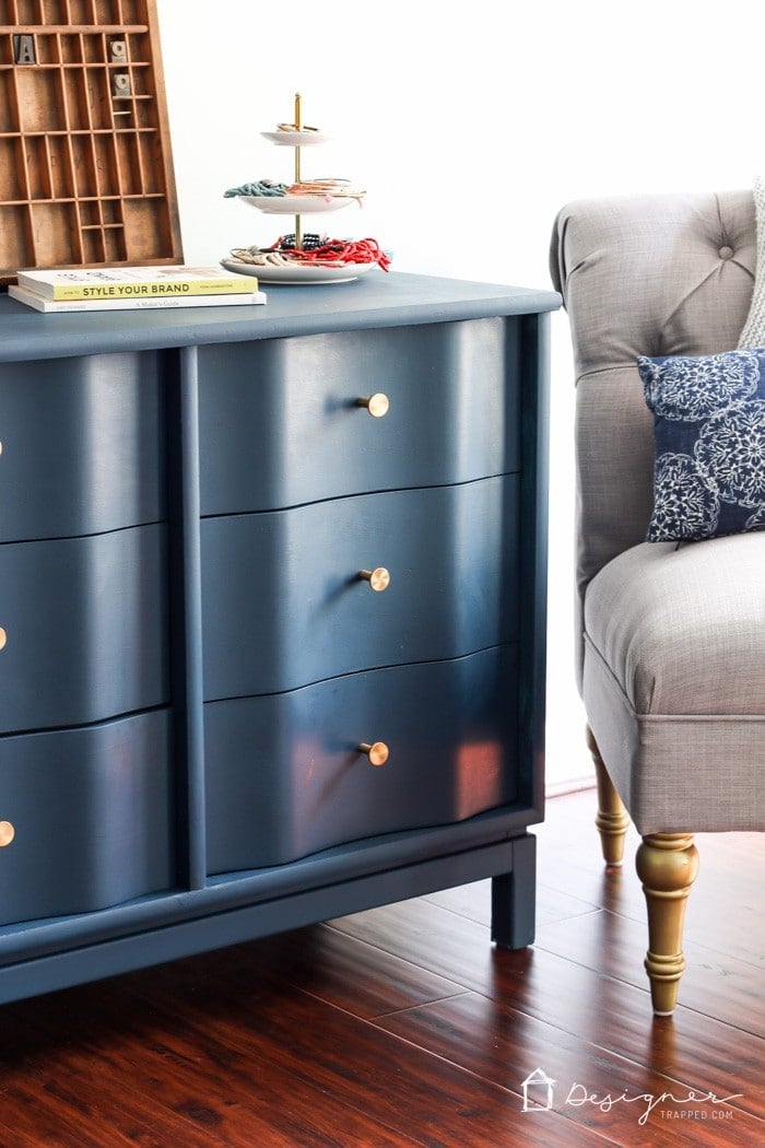 Close-up of a navy blue painted wood dresser with gold knobs, styled with a tiered tray of accessories, books, and a vintage letterpress, placed beside a tufted gray armchair. A polished example of how to paint wood furniture for a modern makeover. Text in the lower right corner says, "DesignerTrapped.com" with a drawing of a house as a logo.