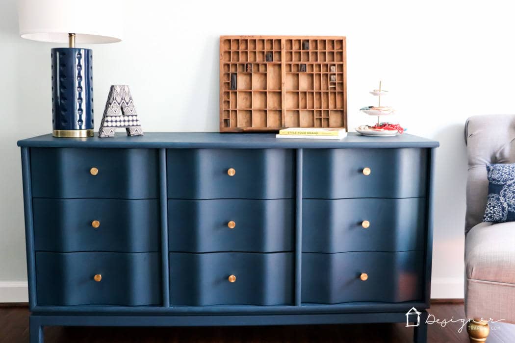 Bold navy blue dresser with gold knobs and a wavy front has a and a smooth finish. Modern decor and a few books sit on top and a gray chair is to the right. A great example of a successful DIY makeover showing how to paint wood furniture for long-lasting results.