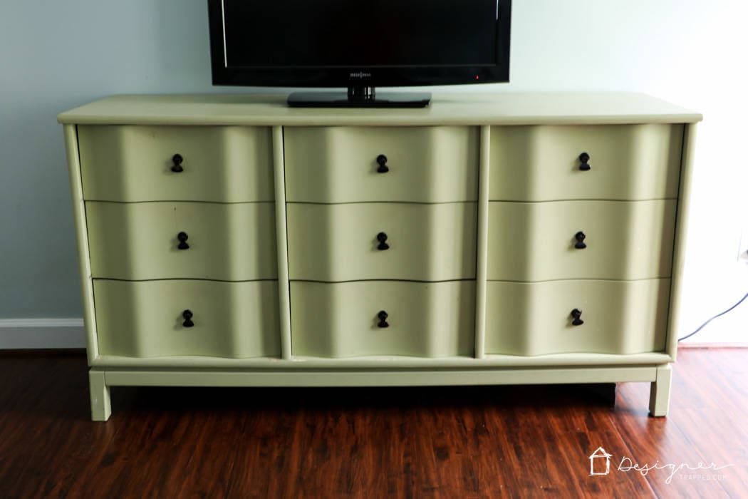 A light green-yellow dresser with curved drawer fronts has a flat screen TV on it and is against a light blue wall on a hardwood floor.