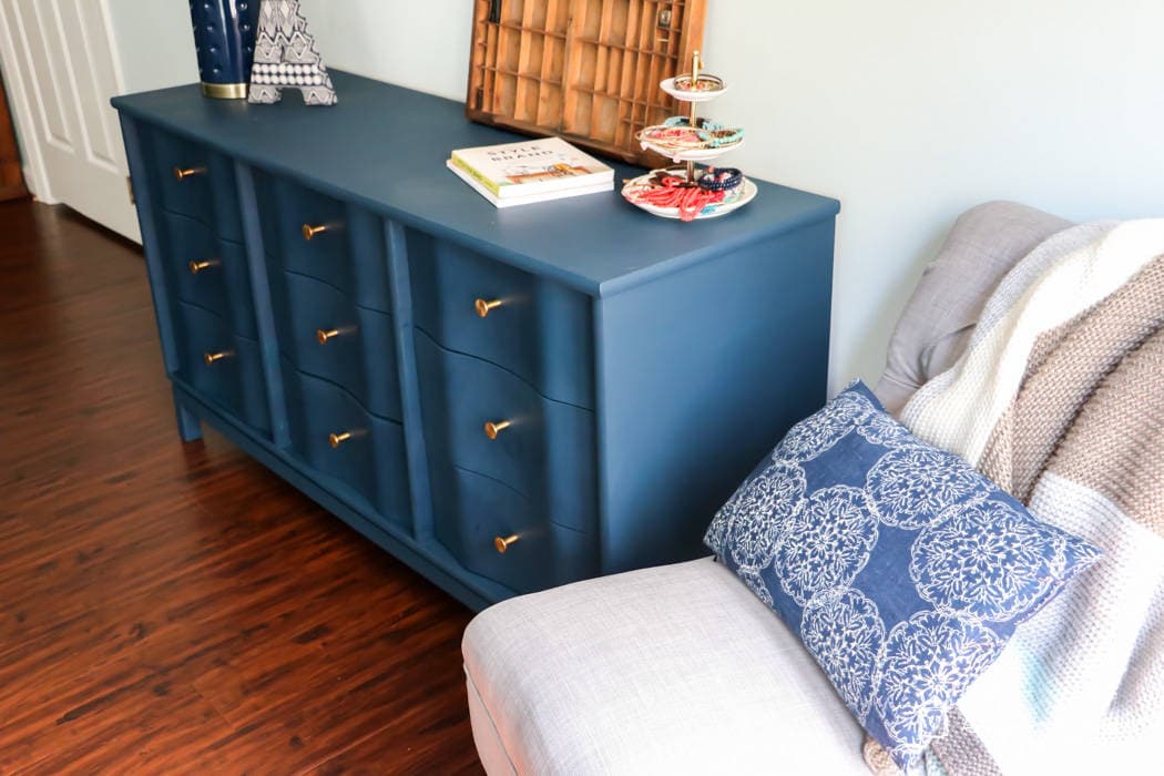 Angled view of a navy-blue painted wood dresser with gold knobs, with a lamp. books, a three-tiered tea and cake holder and more on top. A cozy gray armchair with a blue patterned pillow and knit throw is to the right. The simple house outline logo and the website, "DesignerTrapped.com" is in the lower corner. This dresser is an example of how to paint wood furniture.