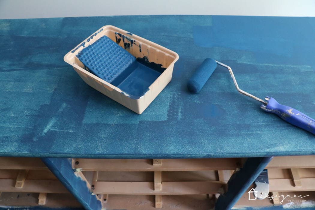 An above view of a dresser being painted blue. A paint tray and roller are on the top of the dresser, and the shelves are empty below. The first coat of blue paint is on the top and shelf supports. The corner text reads, "Designer Trapped.com."