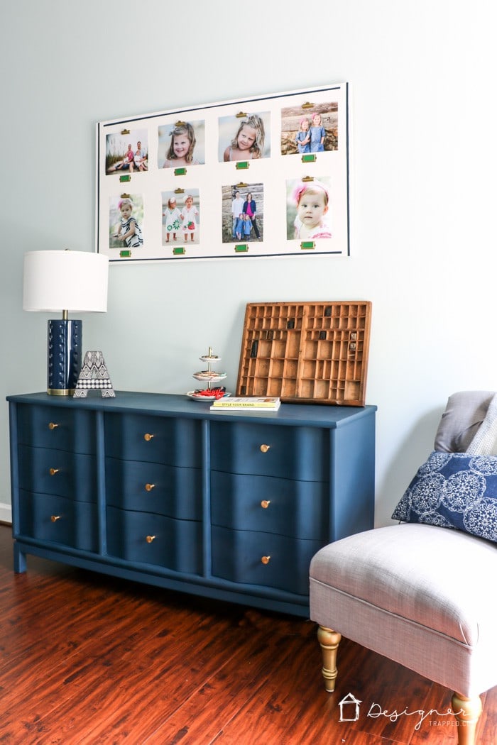 Blue painted dresser with gold knobs next to a gray tufted armchair, topped with decor and books. A large photo collage of children is hanging on the wall above.