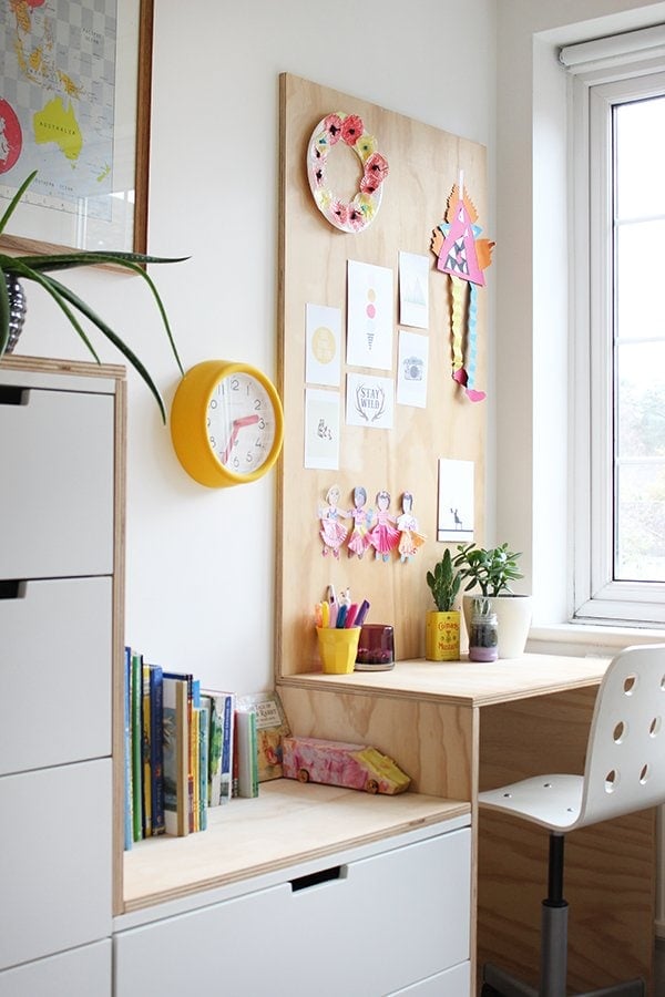 A children's desk and storage corner features a DIY IKEA hack using Nordii drawers and plywood. Children's books, drawings, markers, plants and a yellow clock are dotted along the desk and storage drawers. 