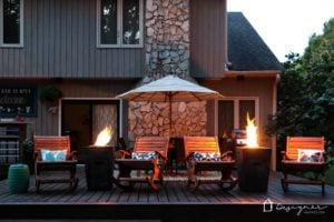 Fire columns on a deck flanking cozy chairs and an umbrella behind a large house.