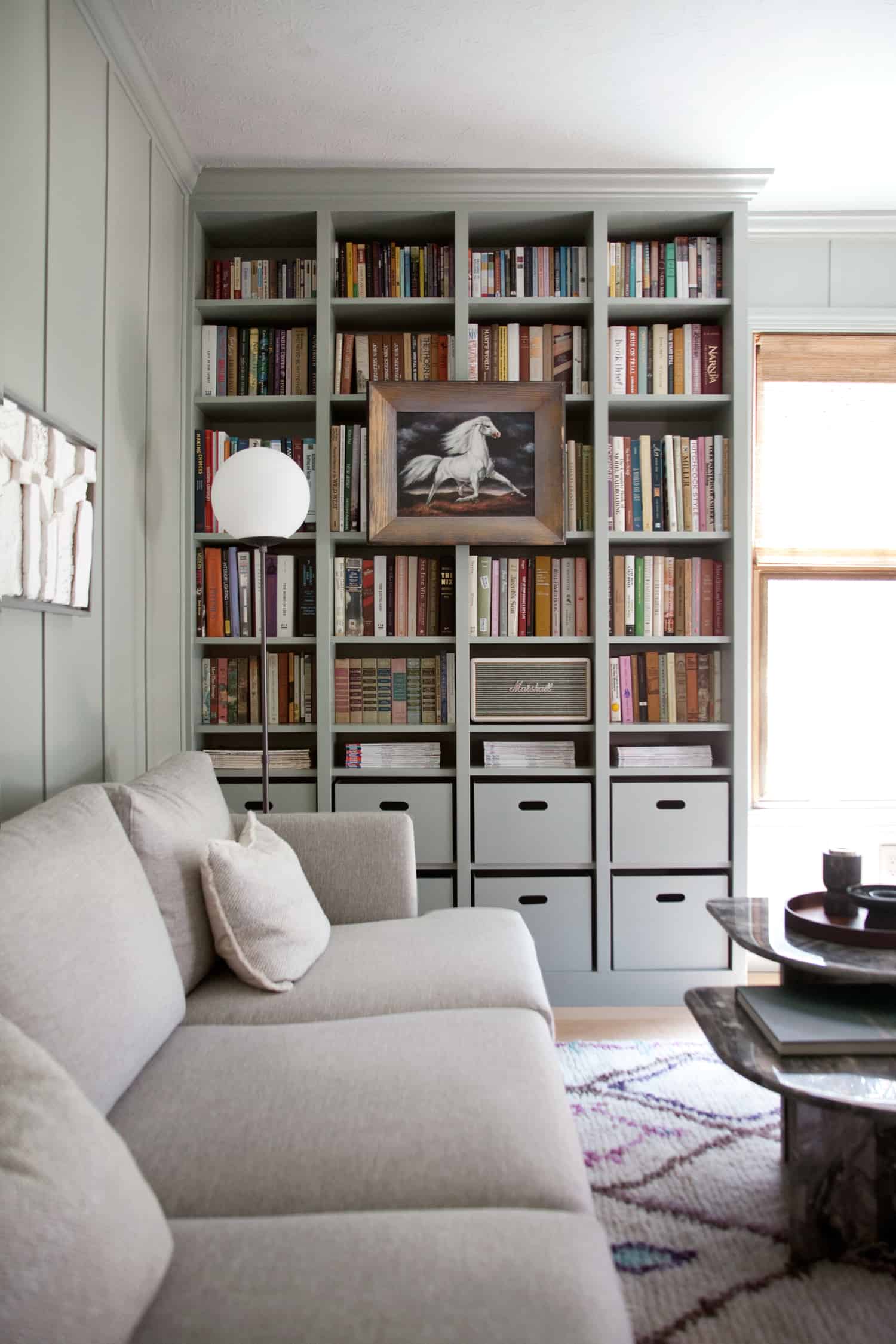 IKEA built-in cabinets in a home library hold floor to ceiling books with a row of cubby boxes on the lower two shelves. A gray couch rests again the connecting wall.