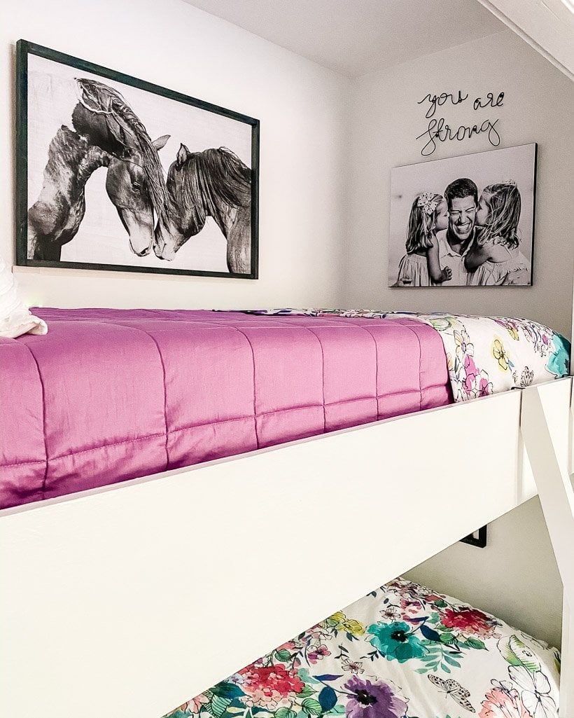 The top of this bedroom's built-in bunk beds has a pink duvet and the wall has a picture of horses, a canvas print of a father with two daughters, and a sign that says, "You are strong."