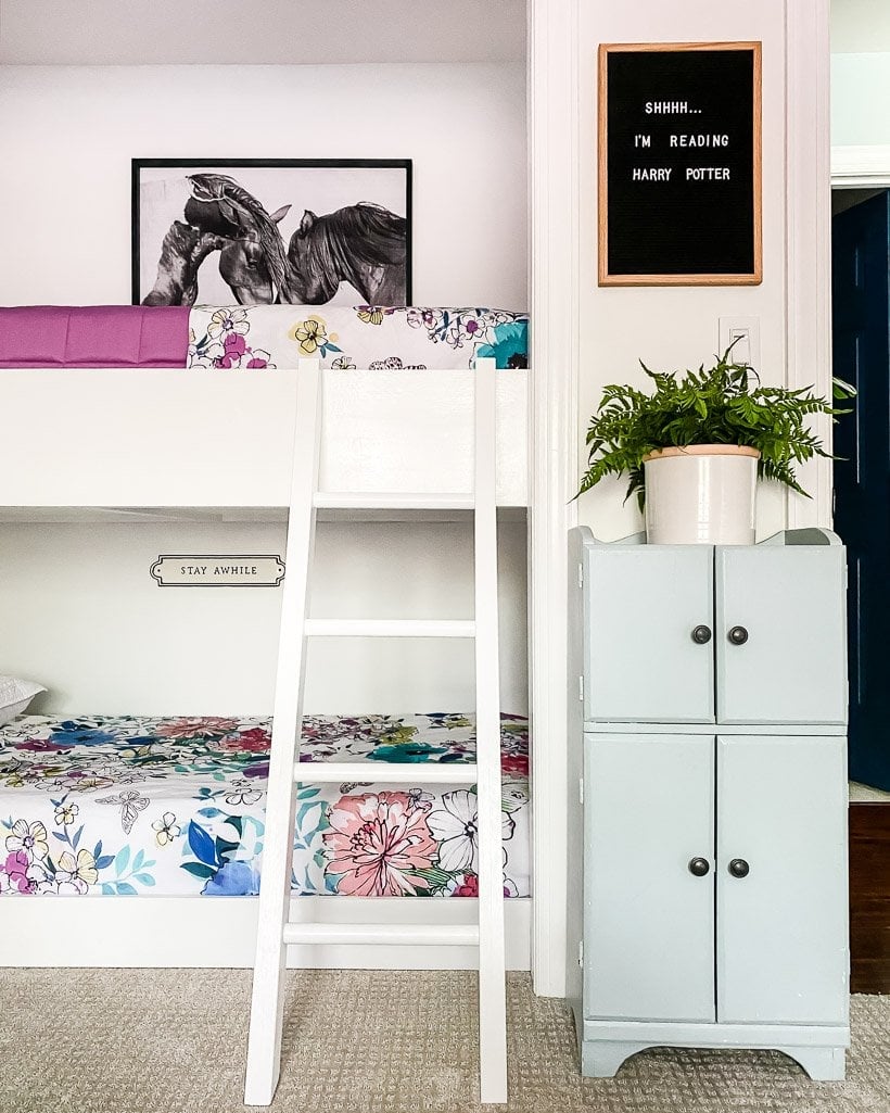 Built-in bunk beds in a girl's bedroom have a white ladder, a tall set of cabinets and a sign that reads, "stay while."