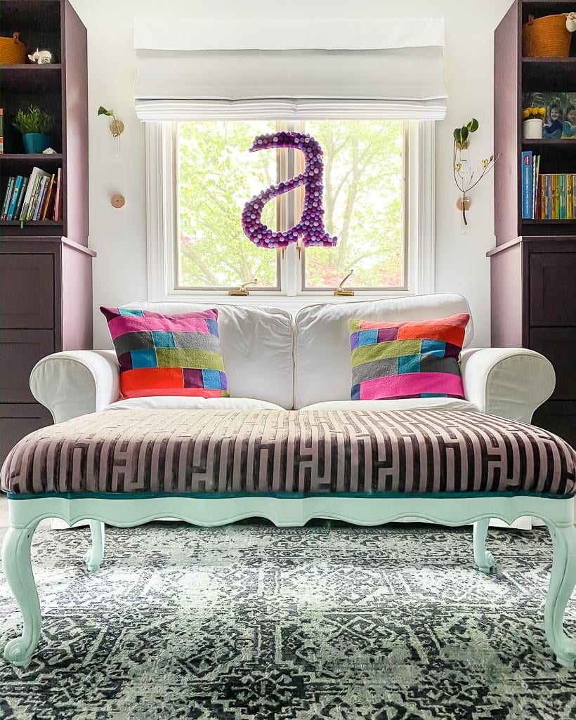 A small-case letter "a" made of purple felt balls hangs in the window of a young girl's bedroom behind a white loveseat. 