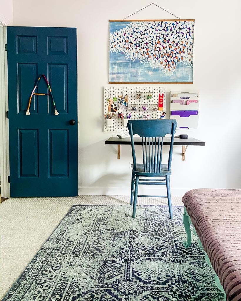 An open blue door to a girl's bedroom featuring a wall-mounted desk, pegboard, and bright hanging artwork. 