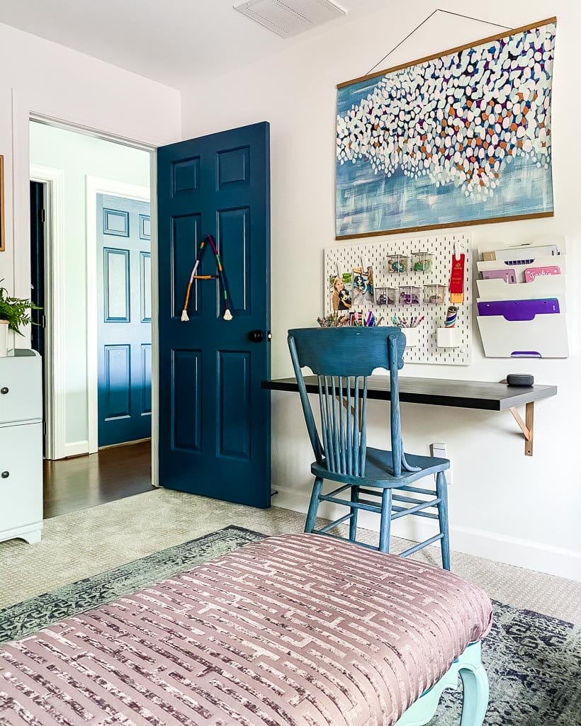 A young girl's bedroom door is painted blue to match the chair in front of a wall-mounted desk, pegboard and large art piece. 