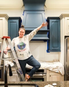 photo of Tasha Agruso of Kaleidoscope Living standing on a ladder in painting clothes in front of partially painted kitchen cabinets