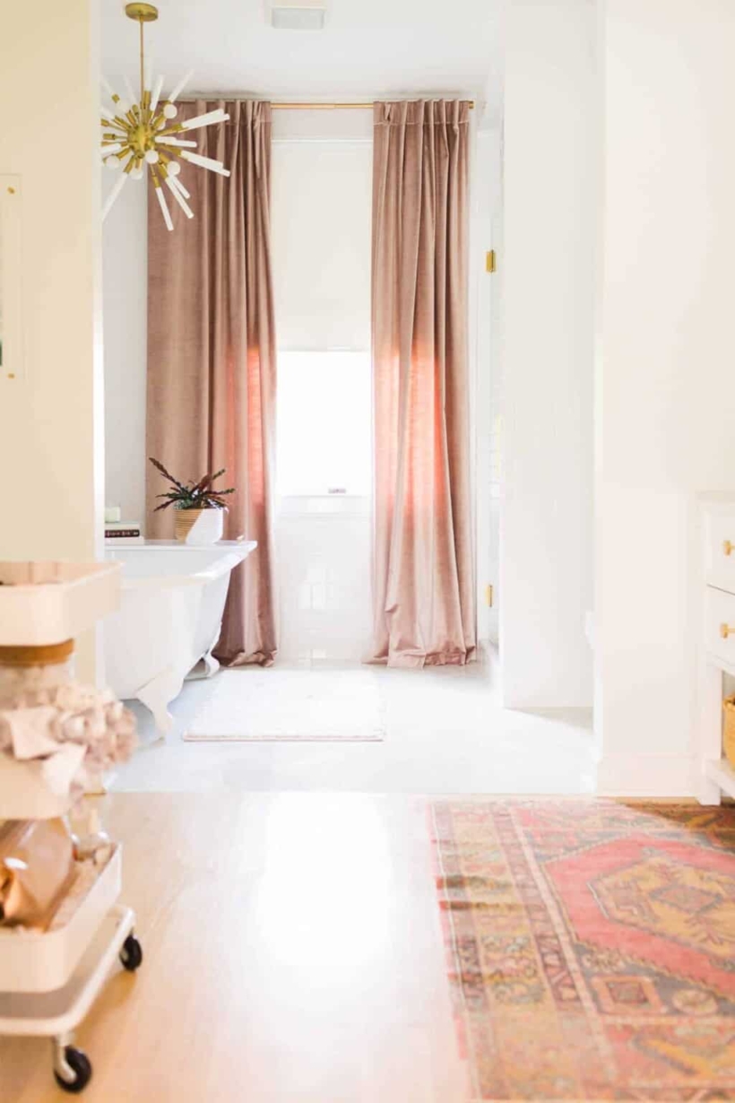 Floor to ceiling curtains frame a sunny window against the back wall of a long primary bathroom. Photo courtesy of "A Beautiful Mess."