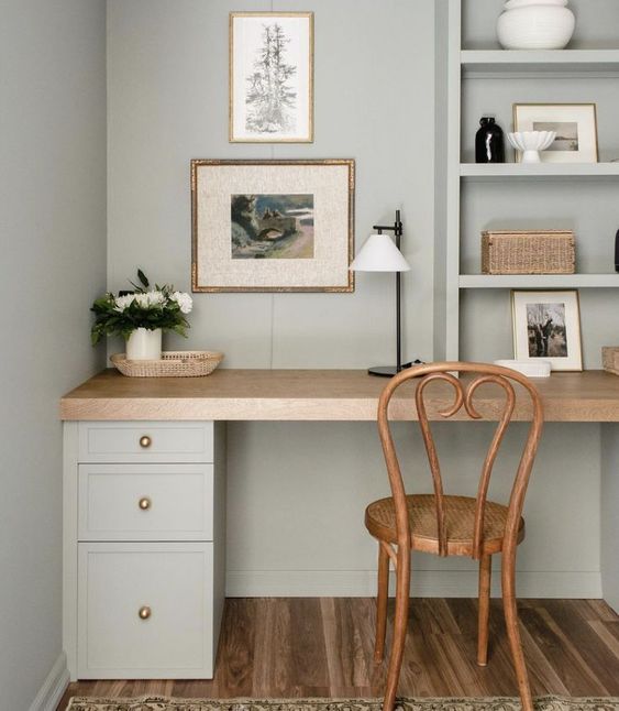 A DIY desk is against a small wall in a light colored room. A wooden chair is in front, and the desk has shelves full of knickknacks, pictures and a plant. This desk uses IKEA Alex drawers and DIY IKEA hacks. Photo from Jyll Mackie. 