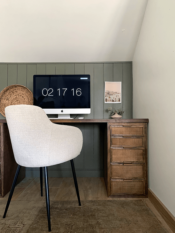 A small desk with a dark stained surface and caned drawers has a computer monitor on top and a modern fabric chair in front. The monitor reads "02 17 16." 