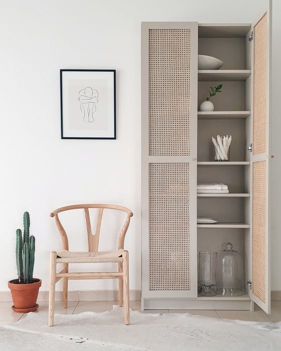 A beige IKEA Billy bookcase has caned doors added to hold extra plates and towels. A simple wooden chair. a framed line drawings, and a cactus are next to the tall cabinet. 