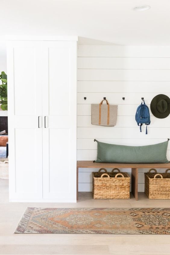 A mudroom setup along the outside wall of a house was made using DIY IKEA hacks like customizing a Pax high cabinet with drawers and a bench with storage underneath. Bags and hats are hung on hooks above the bare bench. 