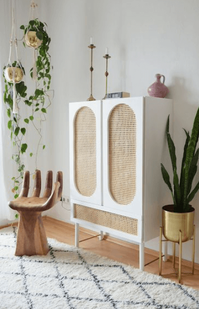 An IKEA rattan Ivar cabinet against a light colored wall, with a large hanging plant in the corner and a wooden hand-shaped chair. 