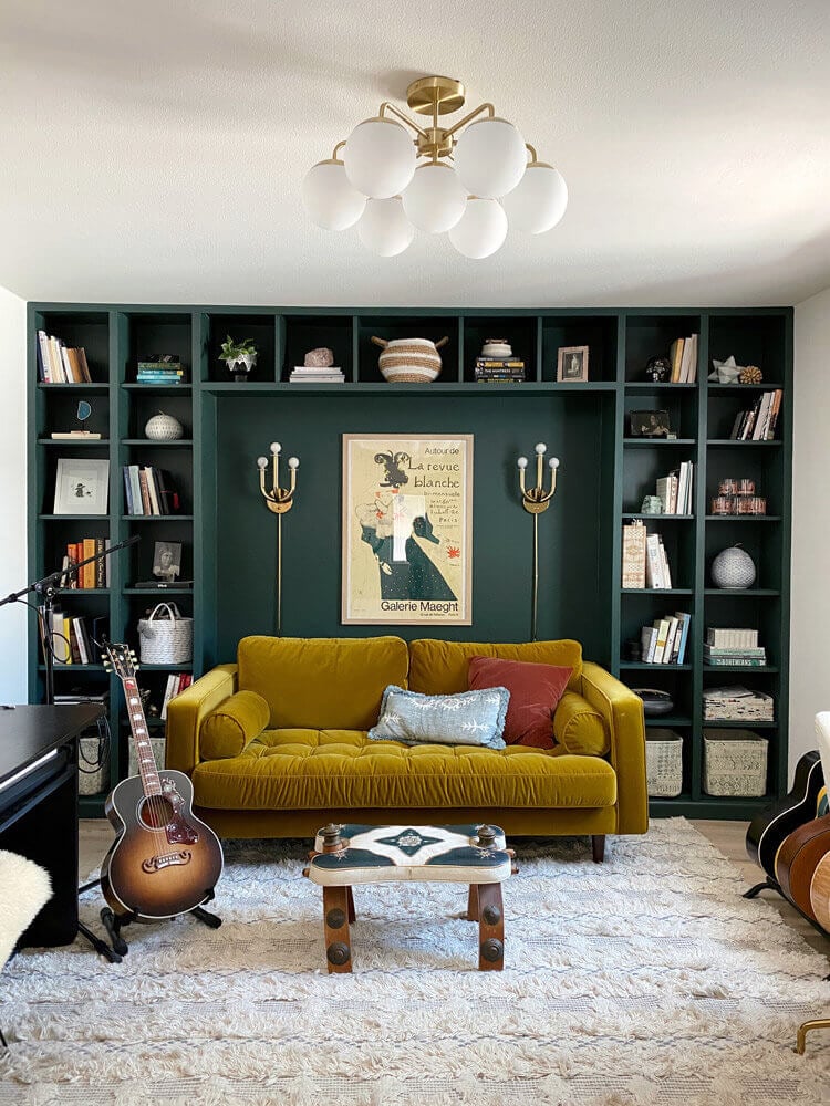 A music room with a dark green wall behind a yellow couch and many guitars. The wall has built-in shelves made from a DIY IKEA hack using Billy bookcases. 