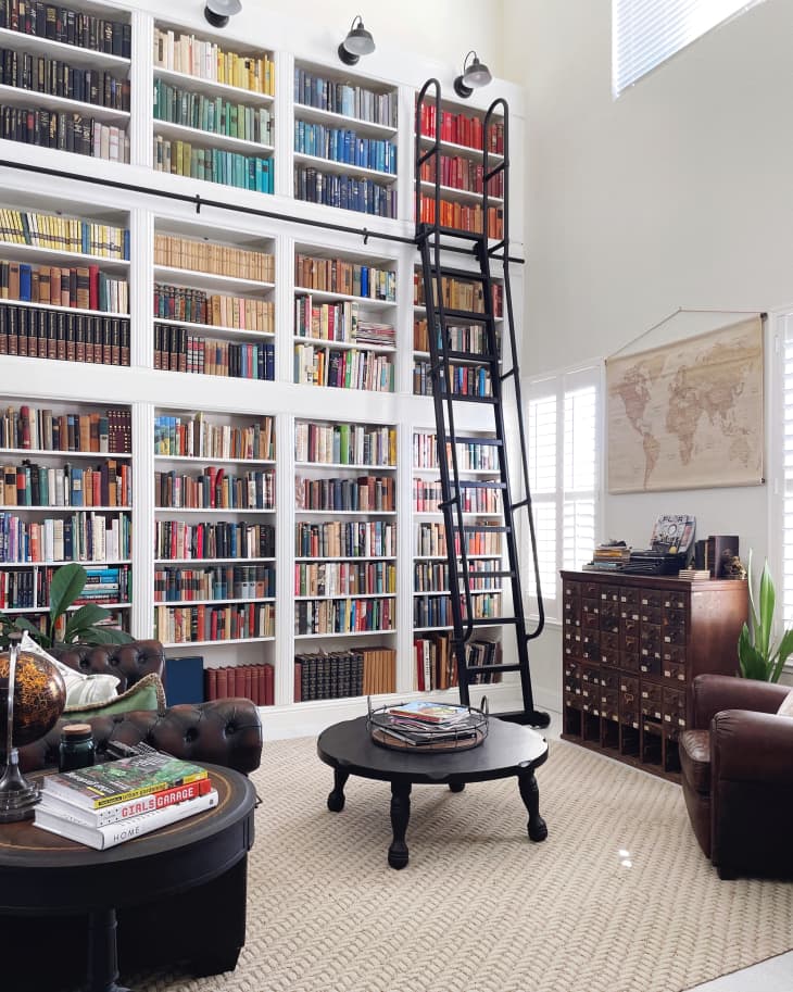 A library in a home with leather chairs, round coffee table, wall map, and an entire backwall of IKEA Billy bookcases converted into a two-story bookshelf, complete with a black iron ladder on wheels. 