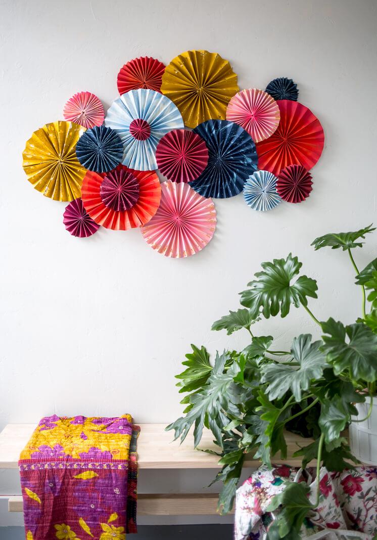 layered fan art made from bright colored card stock and gold paint over a bench with a kantha quilt and plant