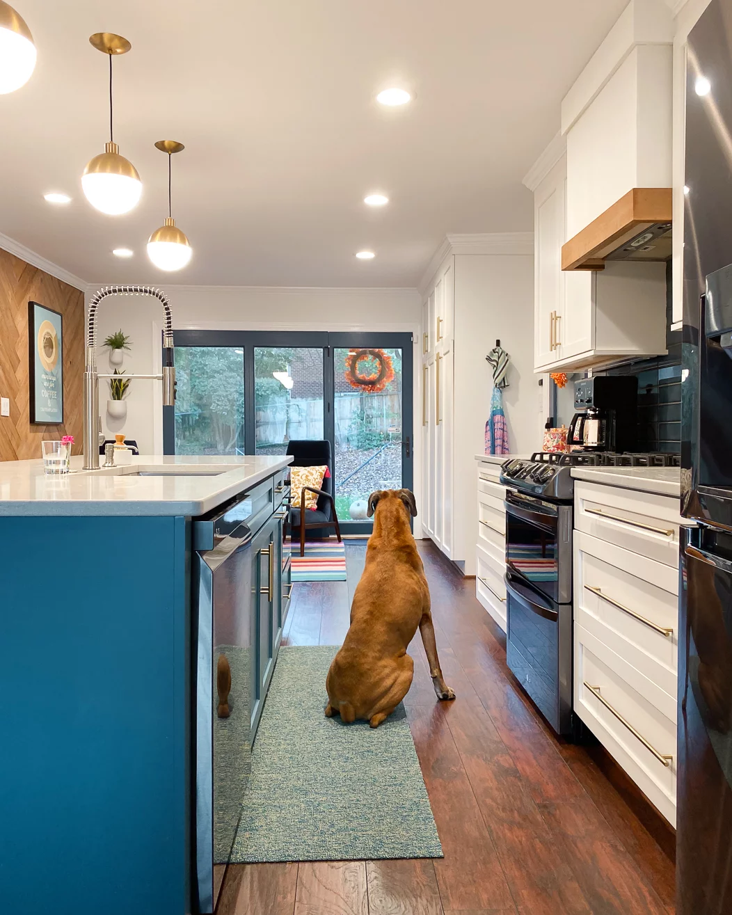 A Chiliewich woven vinyl kitchen rug runner in colorful kitchen with a bright blue center island. A dog sits on the runner looking out sliding glass doors. 