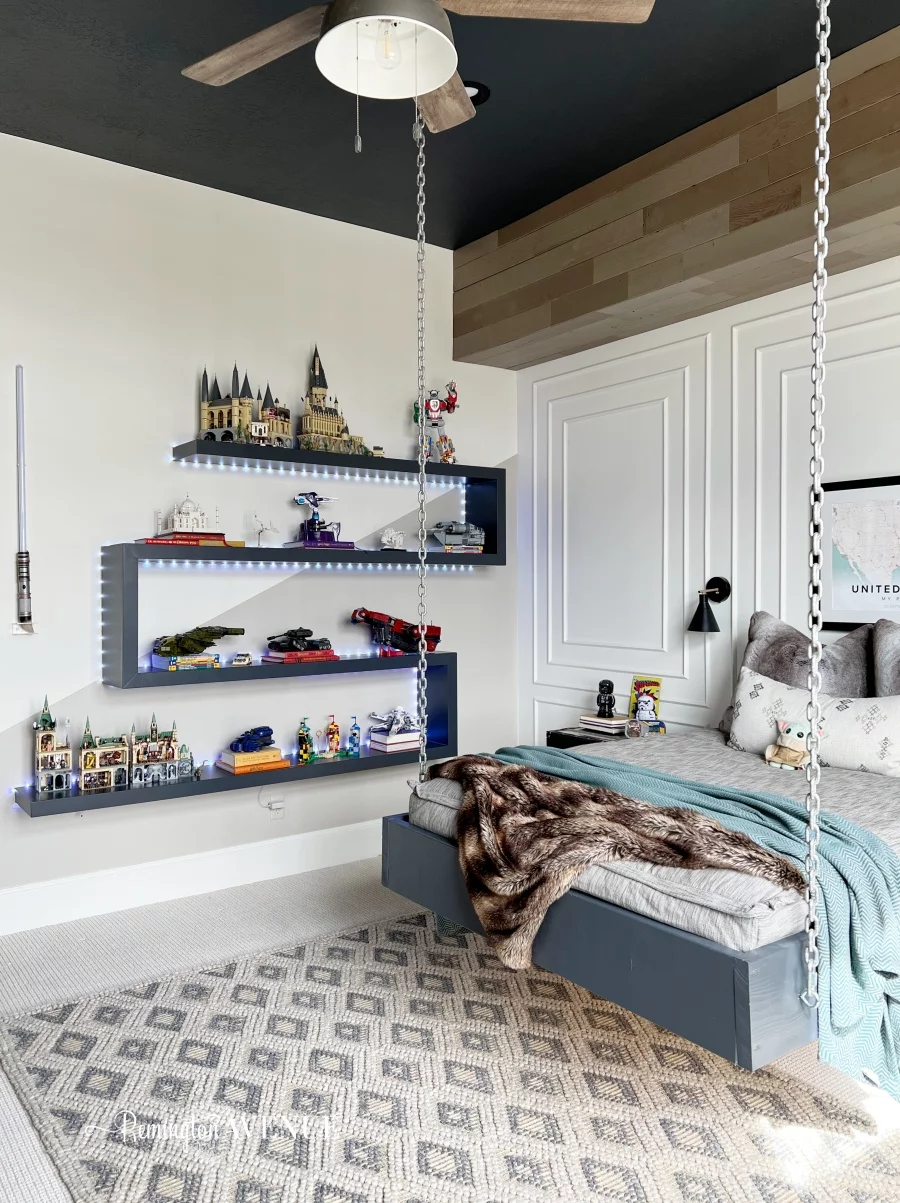 A bed hanging from white chains is suspended over a grey rug. LEGO builds adorn floating zigzag lit shelves. 