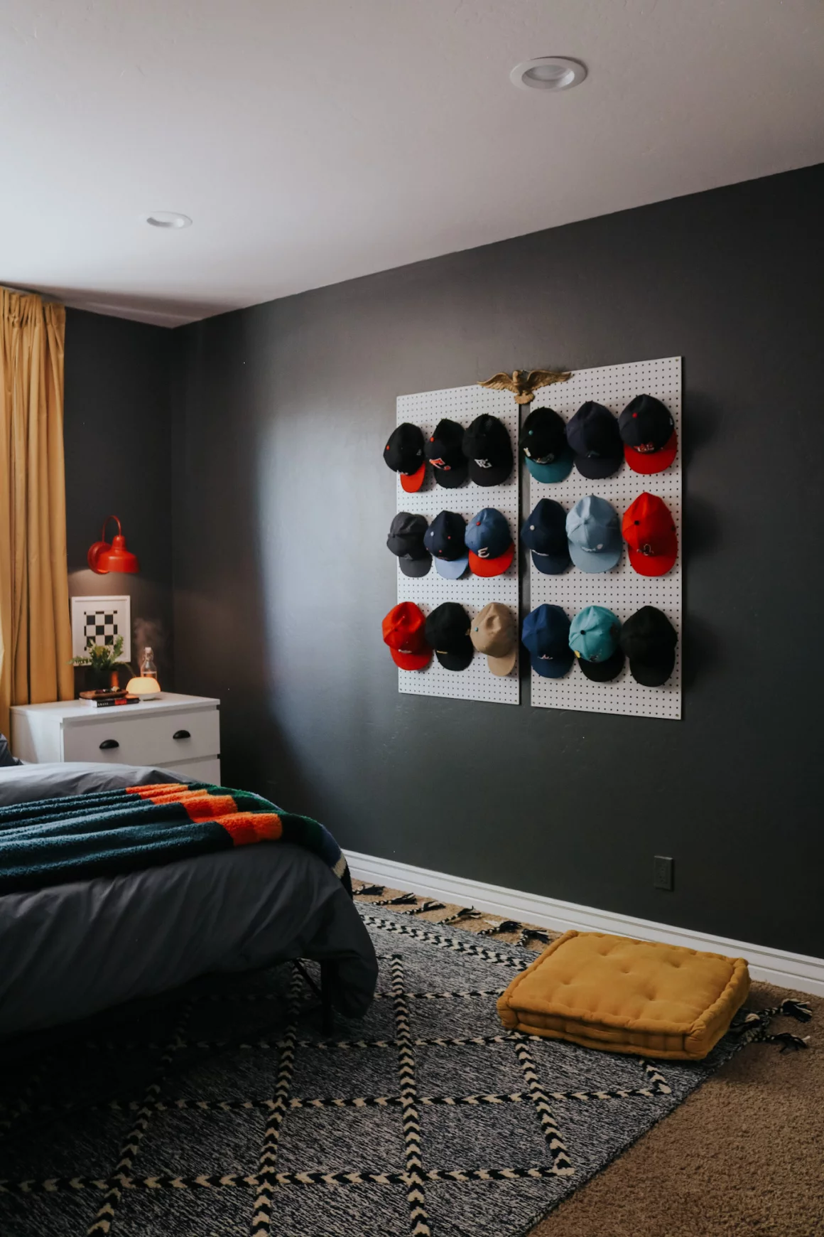 Baseballs hats hanging on pegboard as art on a dark wall with a white nightstand in the corner in a teen boy's room. 