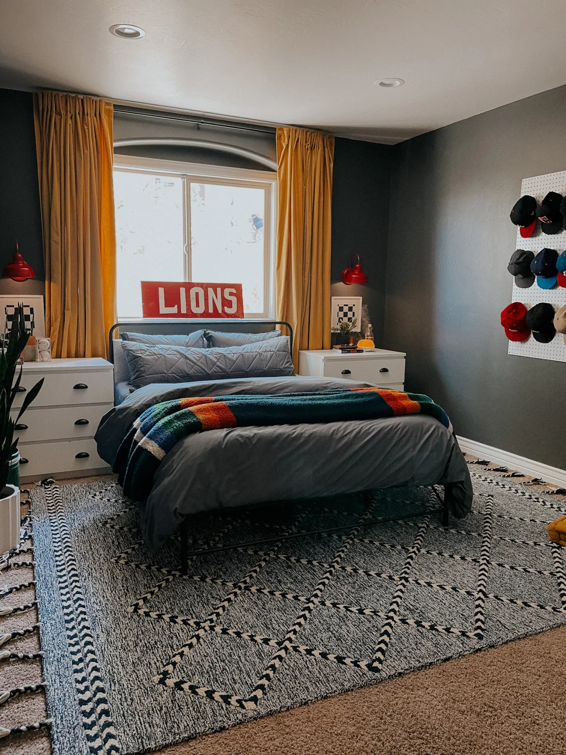 A cozy teen boy bedroom using light and dark colors, white dressers as nightstands, and a pegboard of baseball hats as wall art.