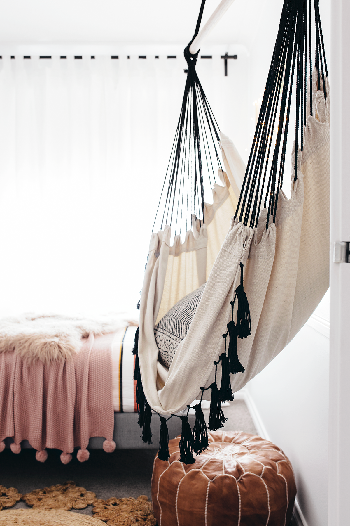 A hanging hammock chair in a teen girl's bedroom with a muted pink comforter on the bed and a leather poof underneath. 