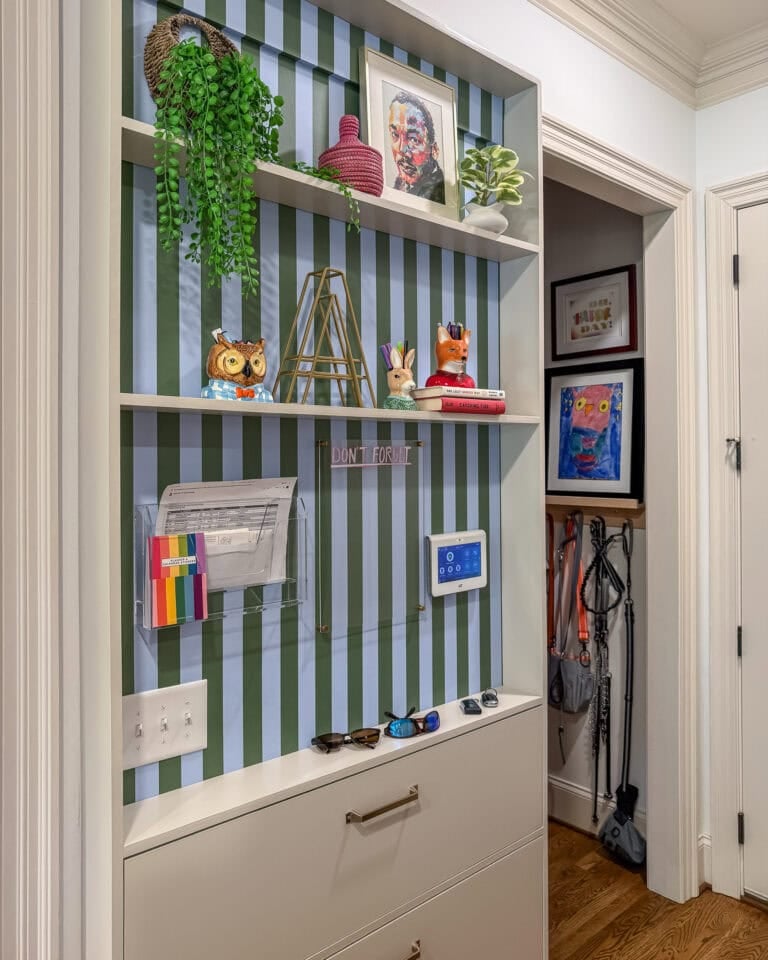 A hanging entryway storage unit with beige drawers and floating shelves set against a green and blue striped wallpaper. The shelves are styled with a family command center and home decor items, including a hanging green plant, a small potted plant, framed art, a small owl figurine, a rabbit figurine, and a fox figurine.