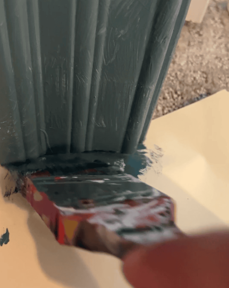 Close-up photo view of paintbrush painting trim along carpeted floor using a manilla file folder under the edge of the trim to protect the carpet from paint